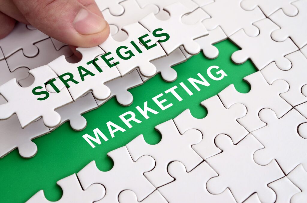 A hand placing a puzzle piece with the word "STRATEGIES" printed in green font to complete a puzzle displaying "MARKETING."